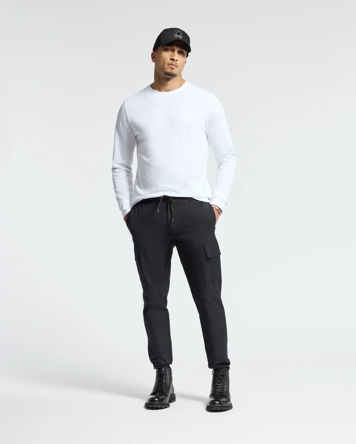 A man stands wearing the Psycho Bunny MENS ALBERTON LONG SLEEVE TEE in white, complemented by a black cap and drawstring black cargo pants. He completes the look with black boots against a plain, light gray background.