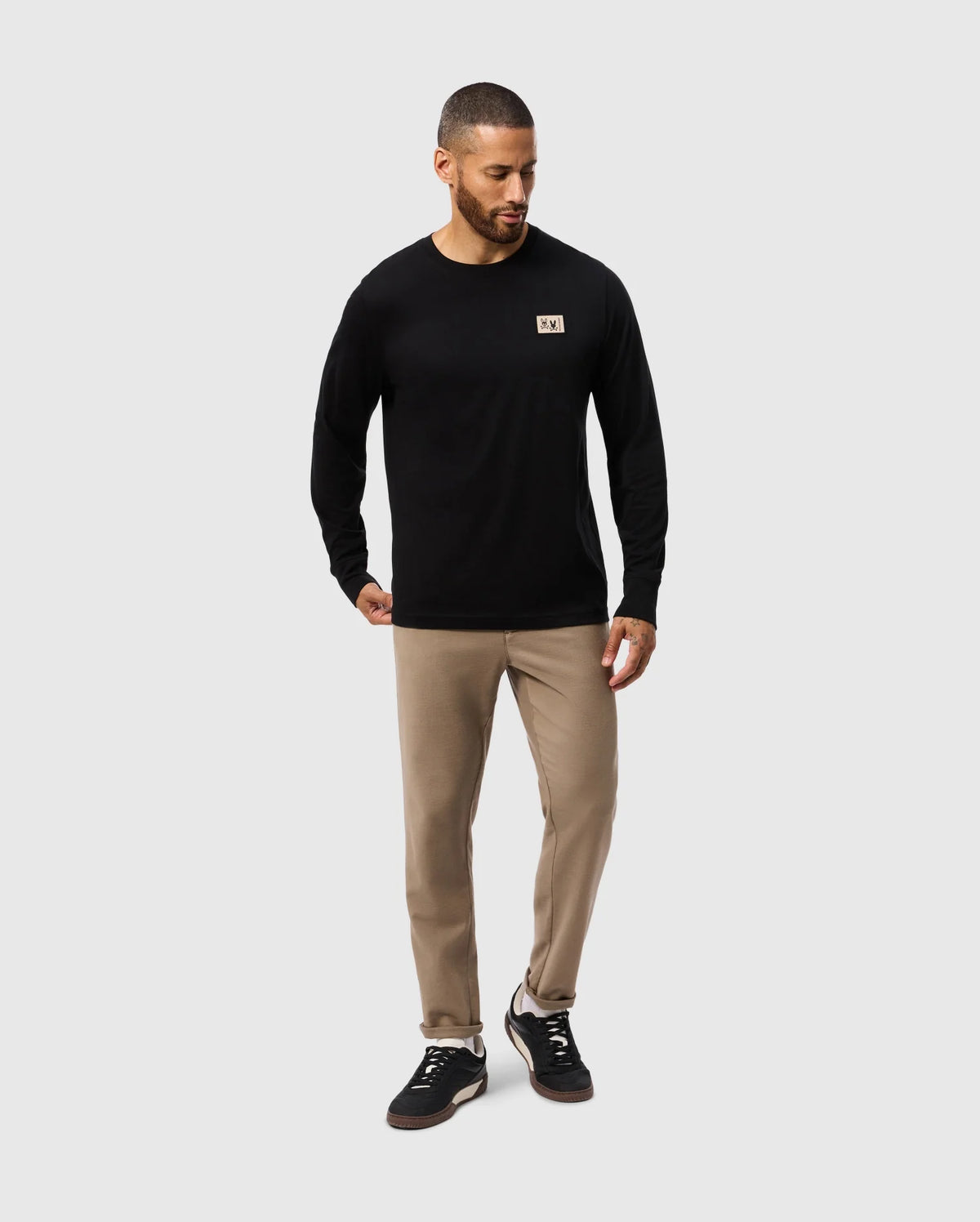 A man in a regular fit, black Psycho Bunny MENS MONROE LONG SLEEVE TEE - B6T975D200, beige pants, and black sneakers stands against a plain white background. Crafted from pima cotton, his shirt enhances the look of his beard and short hair as he gazes slightly to his left.