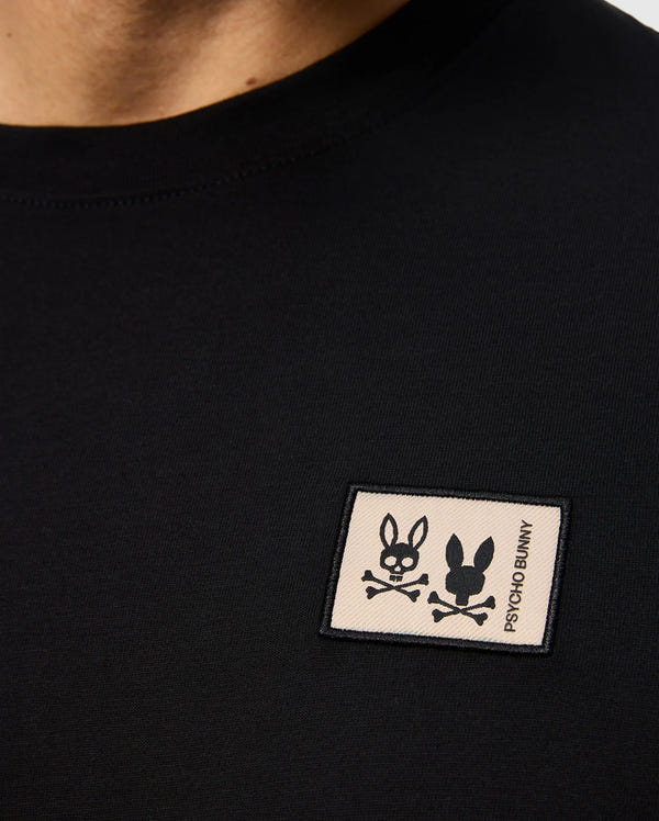 Close-up of the MENS MONROE LONG SLEEVE TEE - B6T975D200, a black shirt made from lightweight pima cotton. It features a small rectangular patch showcasing a playful bunny skull and crossbones design, along with the "Psycho Bunny" text, all crafted for ultimate comfort in a regular fit.