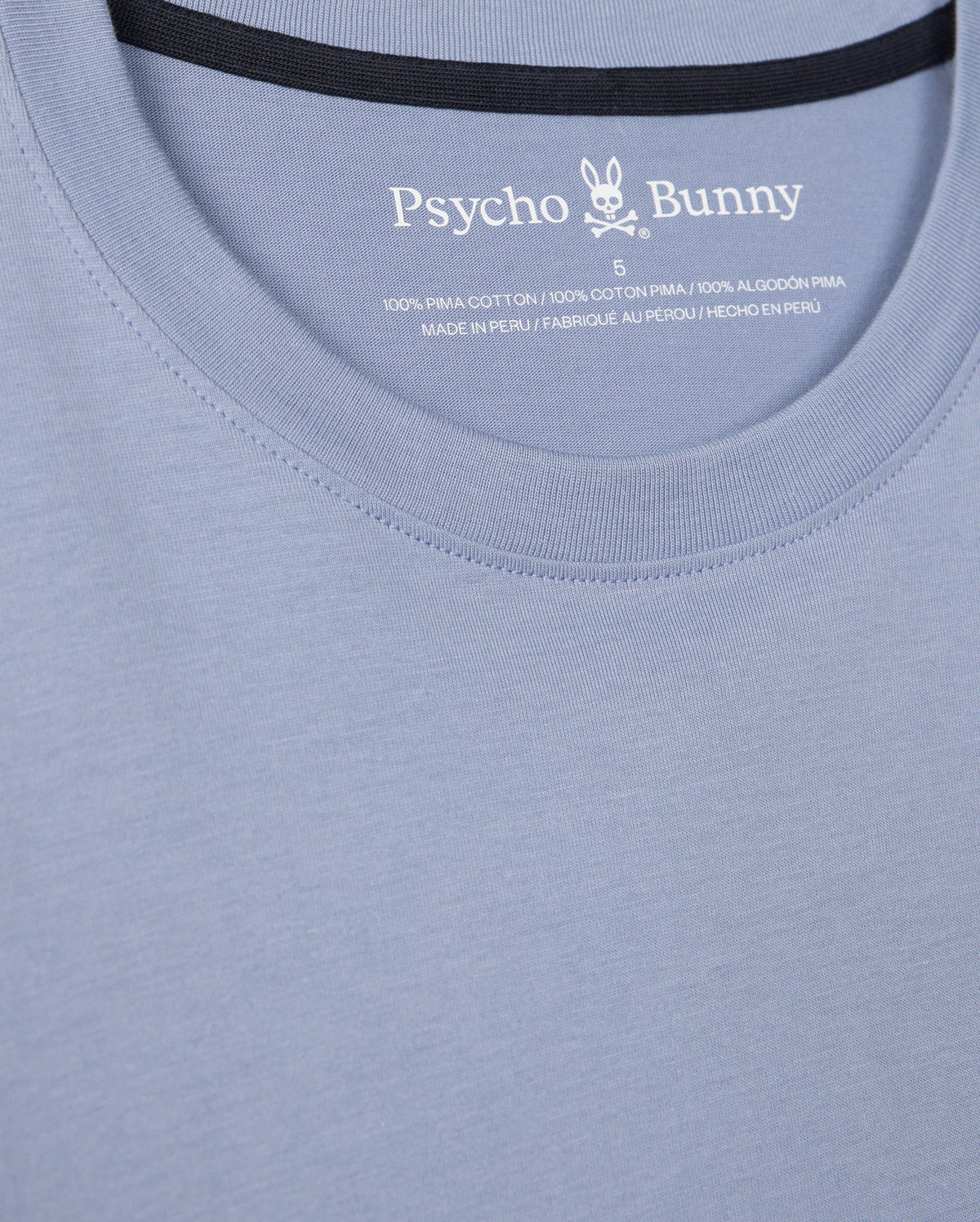 Close-up of a light blue Psycho Bunny Men's Monroe Long Sleeve Tee, highlighting its versatile style. The neckline and upper chest are visible, featuring a label inside the collar that reads "100% Pima Cotton" and "Made in Peru," promising classic comfort and quality.