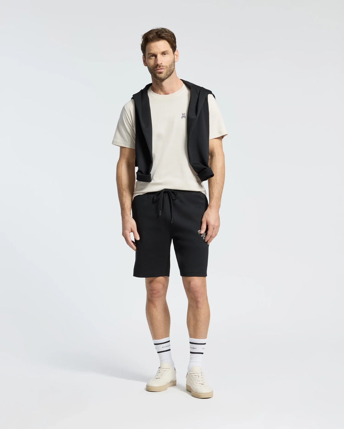 A man stands wearing a Psycho Bunny MENS CLASSIC CREW NECK TEE in white, paired with black shorts and a black jacket tied around his shoulders. He completes the look with striped socks and white sneakers against a plain white background.