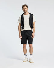 A man stands wearing a Psycho Bunny MENS CLASSIC CREW NECK TEE in white, paired with black shorts and a black jacket tied around his shoulders. He completes the look with striped socks and white sneakers against a plain white background.