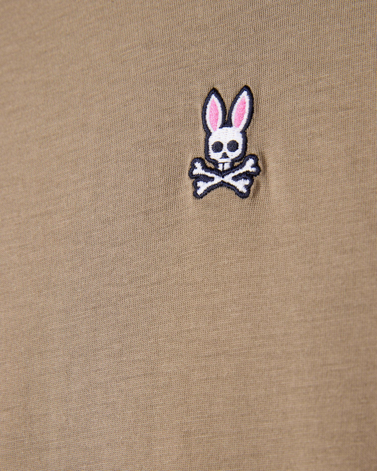 Close-up of the beige fabric made from 100% Pima cotton, showcasing the iconic Psycho Bunny embroidered logo—a white bunny skull with crossbones and pink ears—ideal for the MENS CLASSIC CREW NECK TEE - B6U014CRPC by Psycho Bunny.