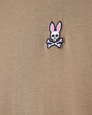Close-up of the beige fabric made from 100% Pima cotton, showcasing the iconic Psycho Bunny embroidered logo—a white bunny skull with crossbones and pink ears—ideal for the MENS CLASSIC CREW NECK TEE - B6U014CRPC by Psycho Bunny.