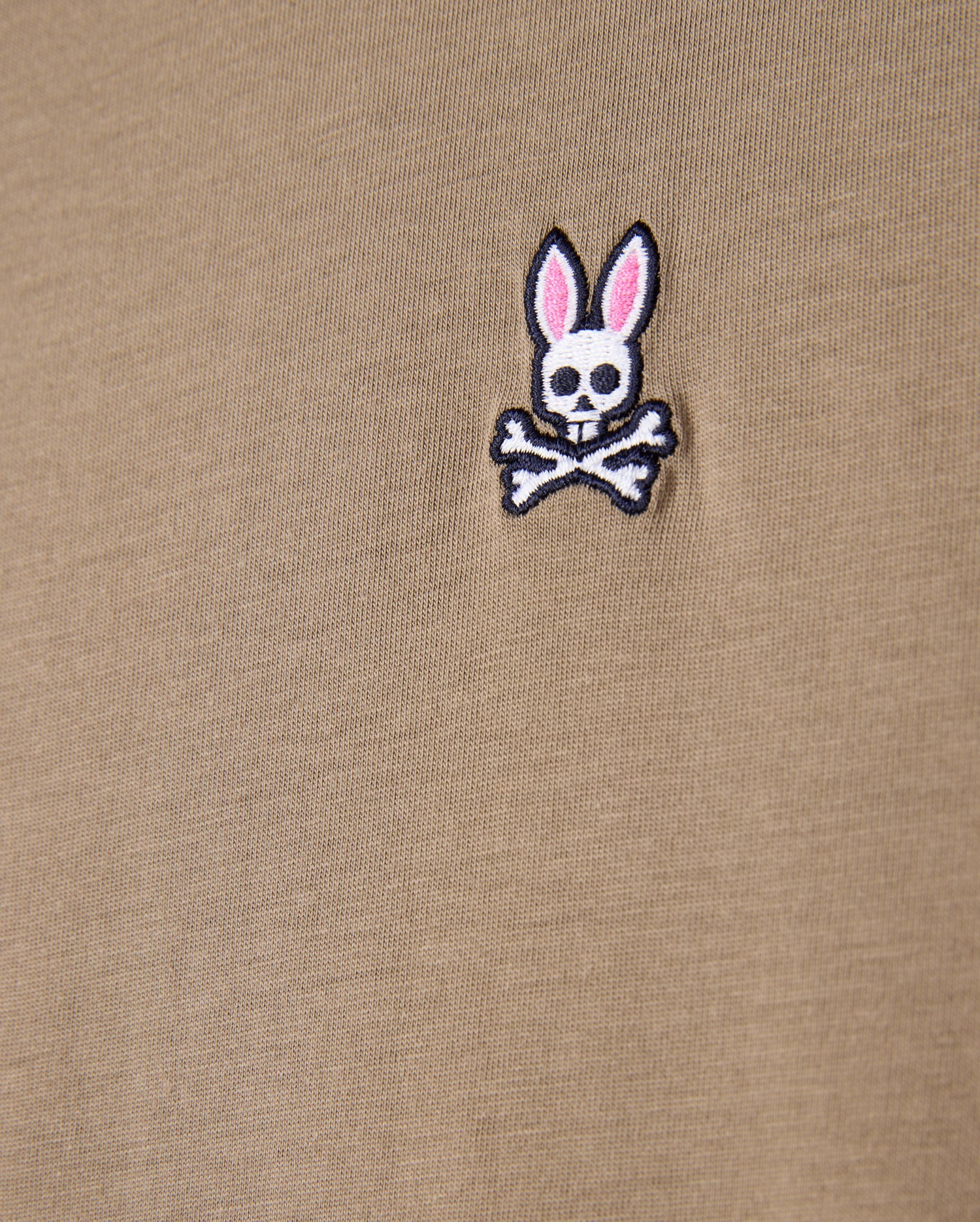 Close-up of the beige fabric made from 100% Pima cotton, showcasing the iconic Psycho Bunny embroidered logo—a white bunny skull with crossbones and pink ears—ideal for the MENS CLASSIC CREW NECK TEE - B6U014CRPC by Psycho Bunny.
