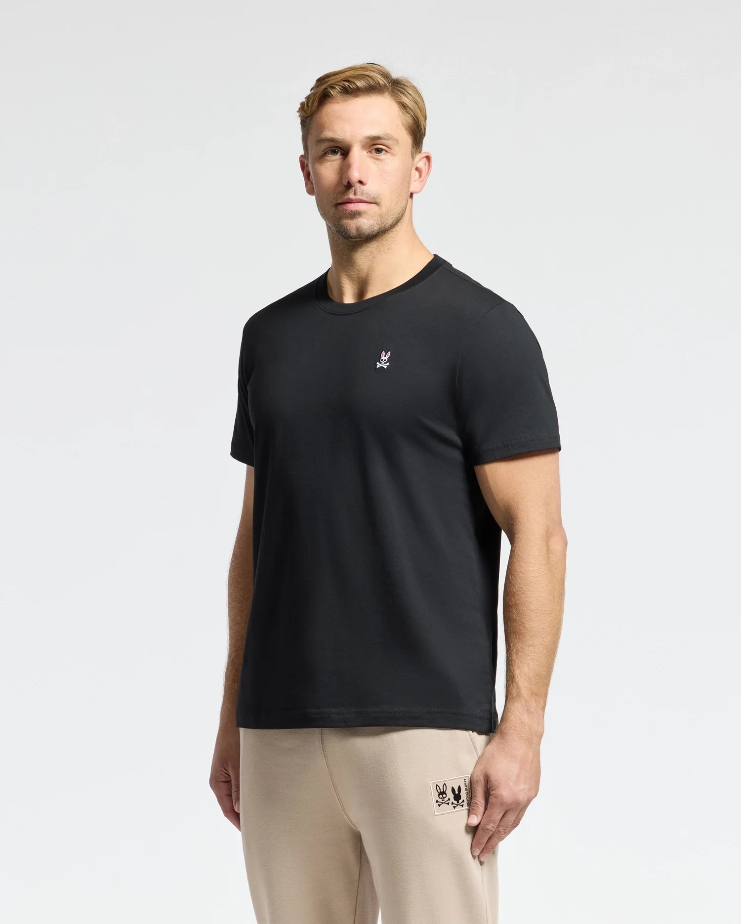 A man stands facing the camera wearing a plain black Psycho Bunny MENS CLASSIC CREW NECK TEE - B6U014CRPC, crafted from 100% Pima cotton, adorned with a small embroidered logo on the left chest, and beige pants. The neutral light gray background accentuates this ideal wardrobe essential.