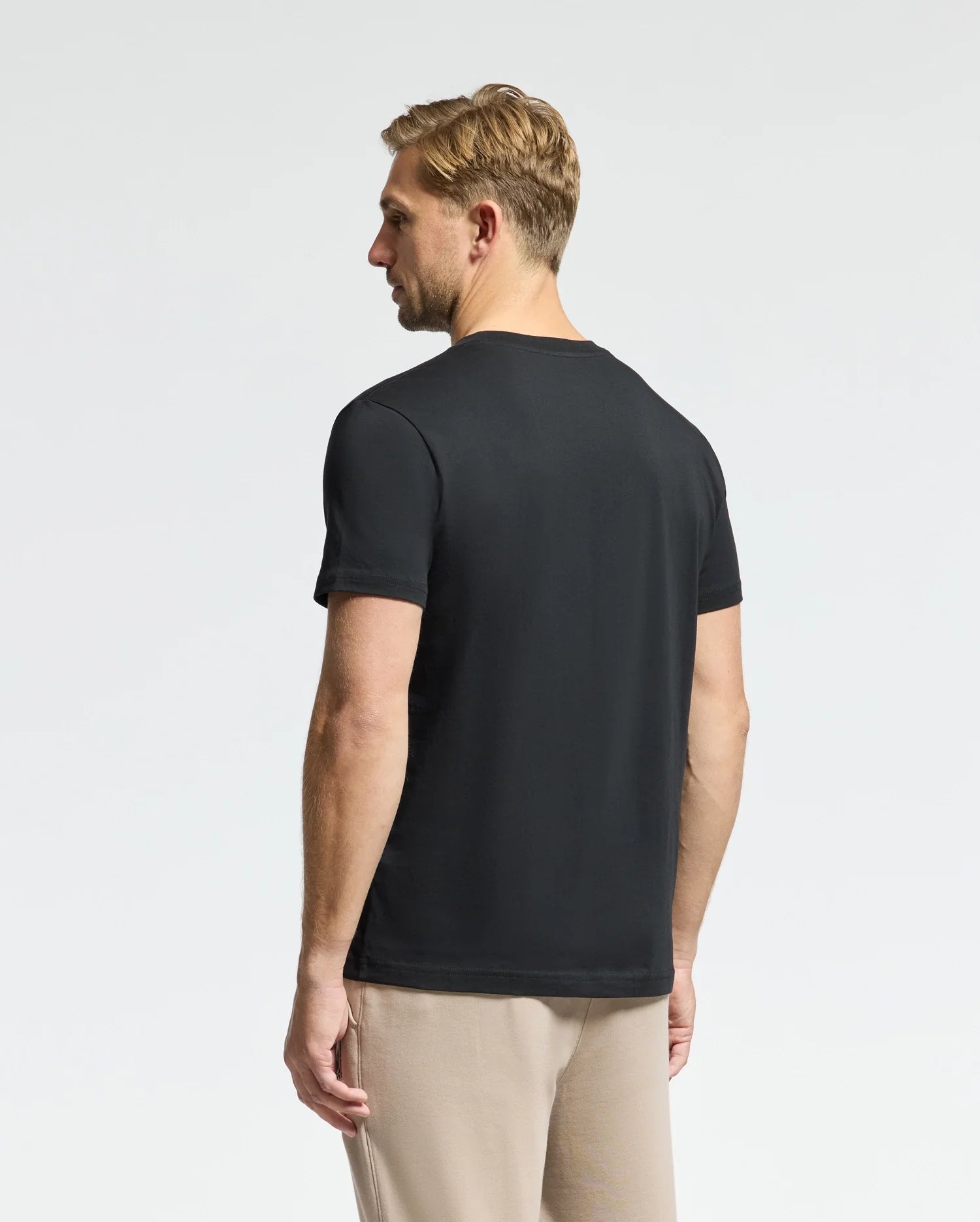 A man with short hair is wearing the Psycho Bunny MENS CLASSIC CREW NECK TEE - B6U014CRPC, a staple in his wardrobe. The black T-shirt, made from 100% Pima cotton for maximum comfort, elegantly complements his beige pants as he stands with his back to the camera against a plain white background.