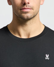 A close-up of a man wearing the Psycho Bunny MENS CLASSIC CREW NECK TEE - B6U014CRPC, crafted from 100% Pima cotton and adorned with a small embroidered logo featuring a bunny and crossed bones on the chest. The image highlights wardrobe essentials with the man's neck and upper chest set against a plain background.
