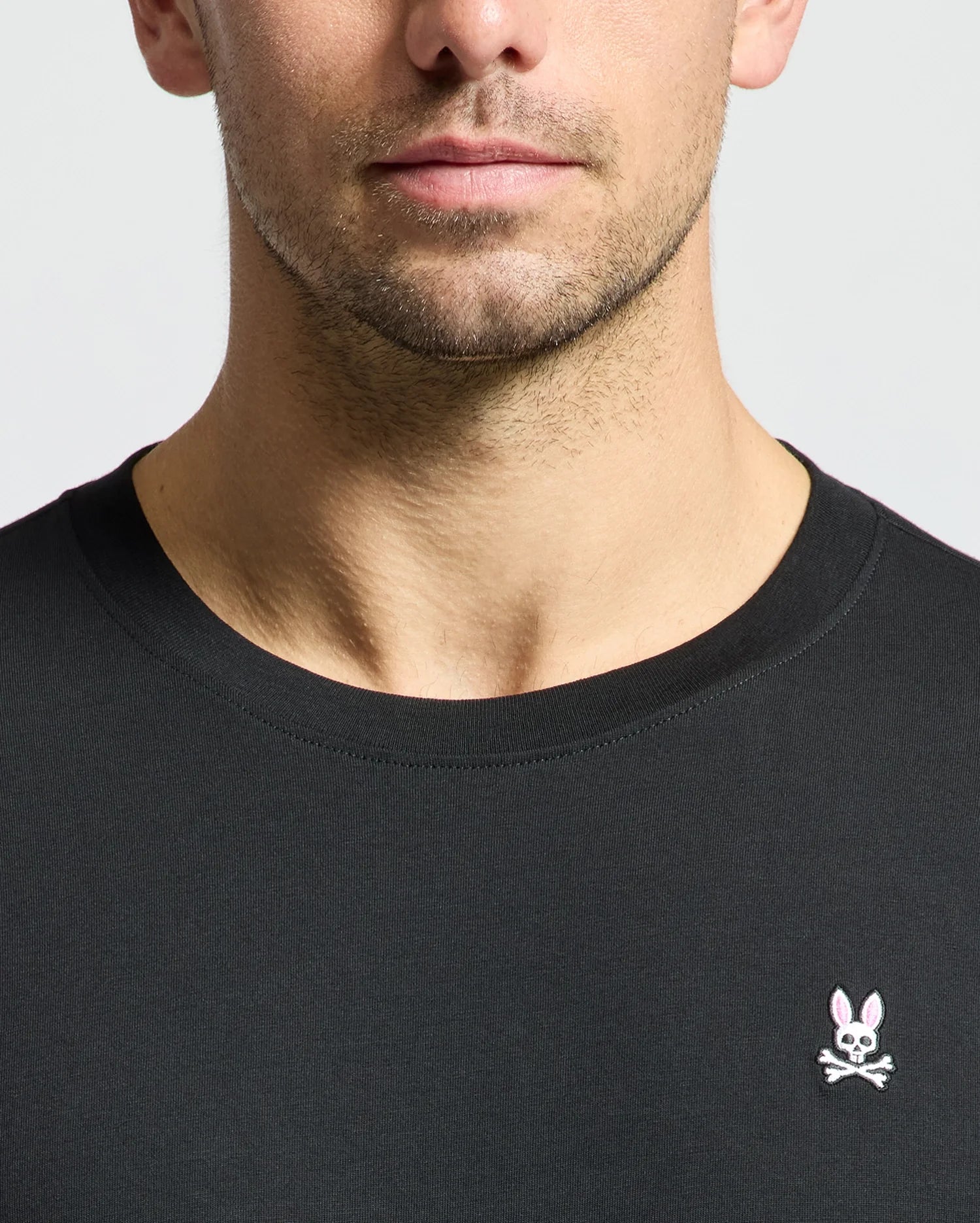 A close-up of a man wearing the Psycho Bunny MENS CLASSIC CREW NECK TEE - B6U014CRPC, crafted from 100% Pima cotton and adorned with a small embroidered logo featuring a bunny and crossed bones on the chest. The image highlights wardrobe essentials with the man's neck and upper chest set against a plain background.