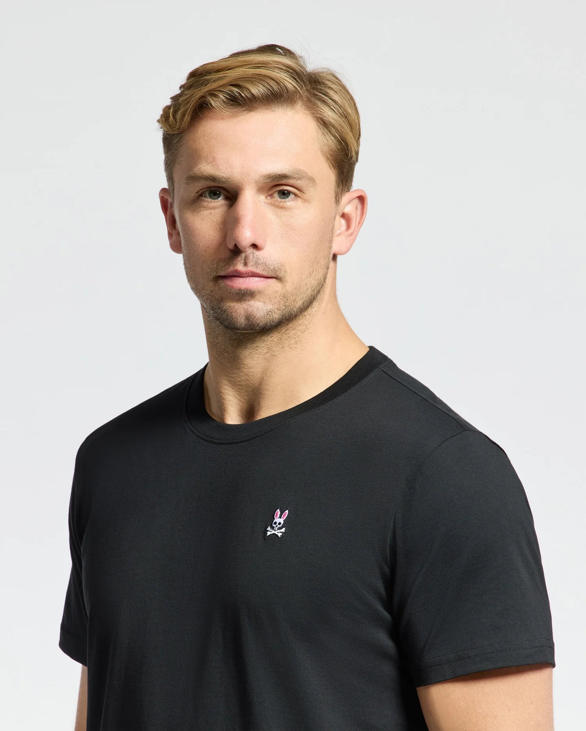 A man with short, blond hair is wearing the Psycho Bunny MENS CLASSIC CREW NECK TEE - B6U014CRPC, crafted from 100% Pima cotton in a classic black hue. The T-shirt features a small embroidered logo in the center. He stands against a plain, light background with a neutral expression, highlighting these timeless wardrobe essentials.