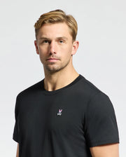 A man with short, blond hair is wearing the Psycho Bunny MENS CLASSIC CREW NECK TEE - B6U014CRPC, crafted from 100% Pima cotton in a classic black hue. The T-shirt features a small embroidered logo in the center. He stands against a plain, light background with a neutral expression, highlighting these timeless wardrobe essentials.