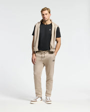 A man stands against a plain background wearing the MENS CLASSIC CREW NECK TEE by Psycho Bunny, made from 100% Pima cotton, paired with beige sweatpants and white sneakers. A beige hoodie is elegantly draped over his shoulders, showcasing essential wardrobe basics.