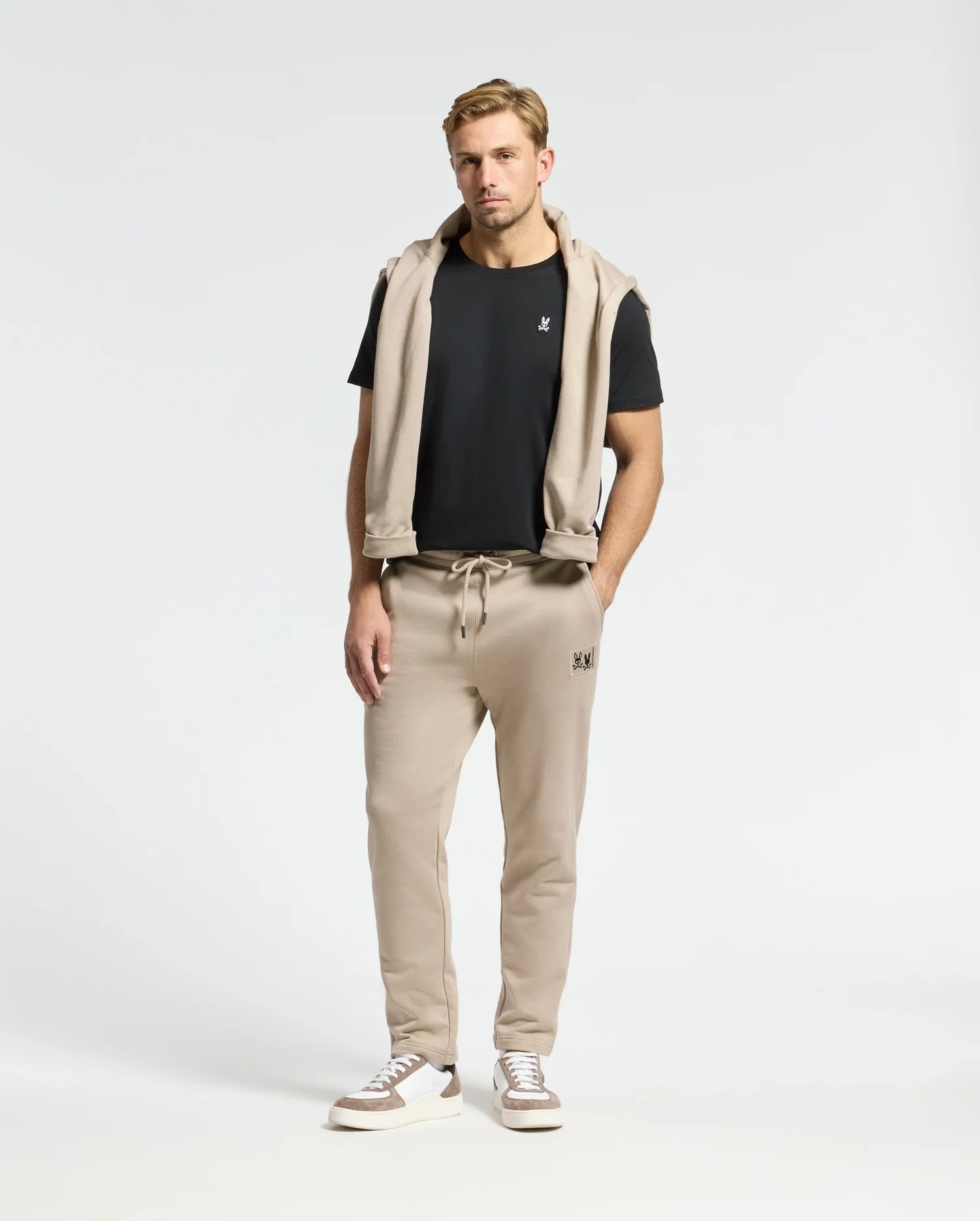 A man stands against a plain background wearing the MENS CLASSIC CREW NECK TEE by Psycho Bunny, made from 100% Pima cotton, paired with beige sweatpants and white sneakers. A beige hoodie is elegantly draped over his shoulders, showcasing essential wardrobe basics.