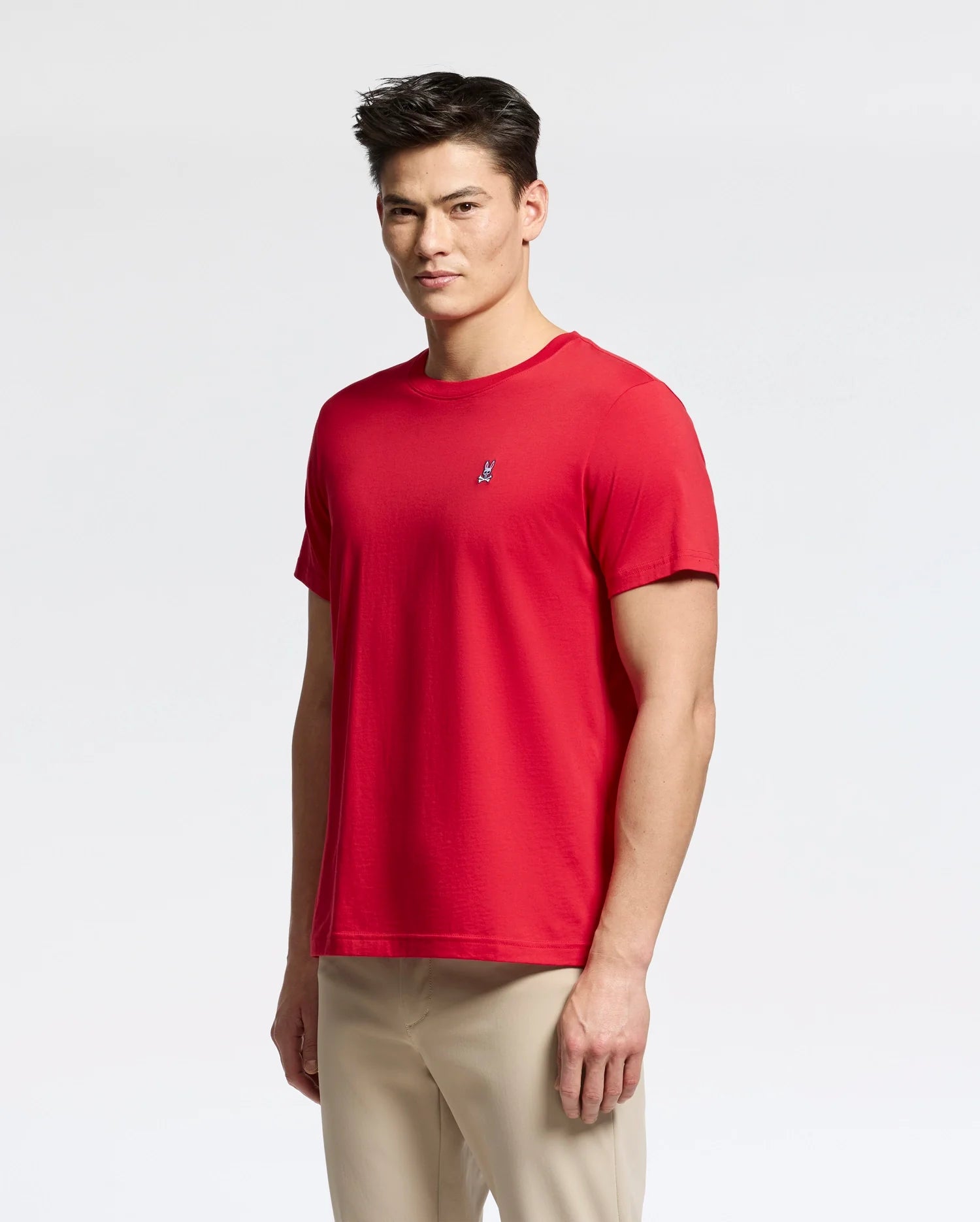 Against a light gray background, a person with short dark hair and beige pants wears a red Psycho Bunny MENS CLASSIC CREW NECK TEE (B6U014CRPC) made from 100% Pima cotton, featuring an embroidered logo on the left chest.