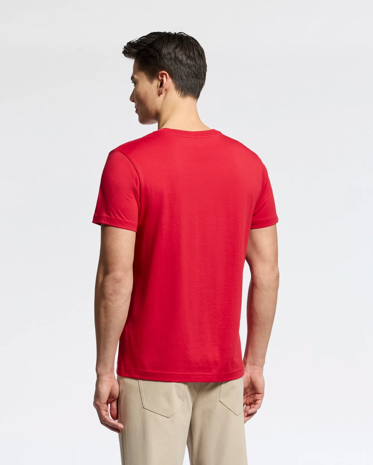 A person with short dark hair stands with their back to the camera, wearing a bright red MENS CLASSIC CREW NECK TEE - B6U014CRPC by Psycho Bunny and beige pants. The tee showcases the embroidered logo on 100% Pima cotton fabric against a plain white background, highlighting its vibrant hue.