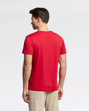 A person with short dark hair stands with their back to the camera, wearing a bright red MENS CLASSIC CREW NECK TEE - B6U014CRPC by Psycho Bunny and beige pants. The tee showcases the embroidered logo on 100% Pima cotton fabric against a plain white background, highlighting its vibrant hue.