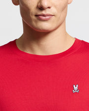 A person wearing a Psycho Bunny men's classic crew neck tee (B6U014CRPC) in plain red, made from 100% Pima cotton, with a small embroidered rabbit and crossbones logo.