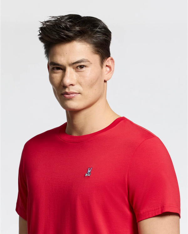 A man wears a MENS CLASSIC CREW NECK TEE - B6U014CRPC from Psycho Bunny, in bright red, made of 100% Pima cotton with a small embroidered logo on the chest, set against a white background.