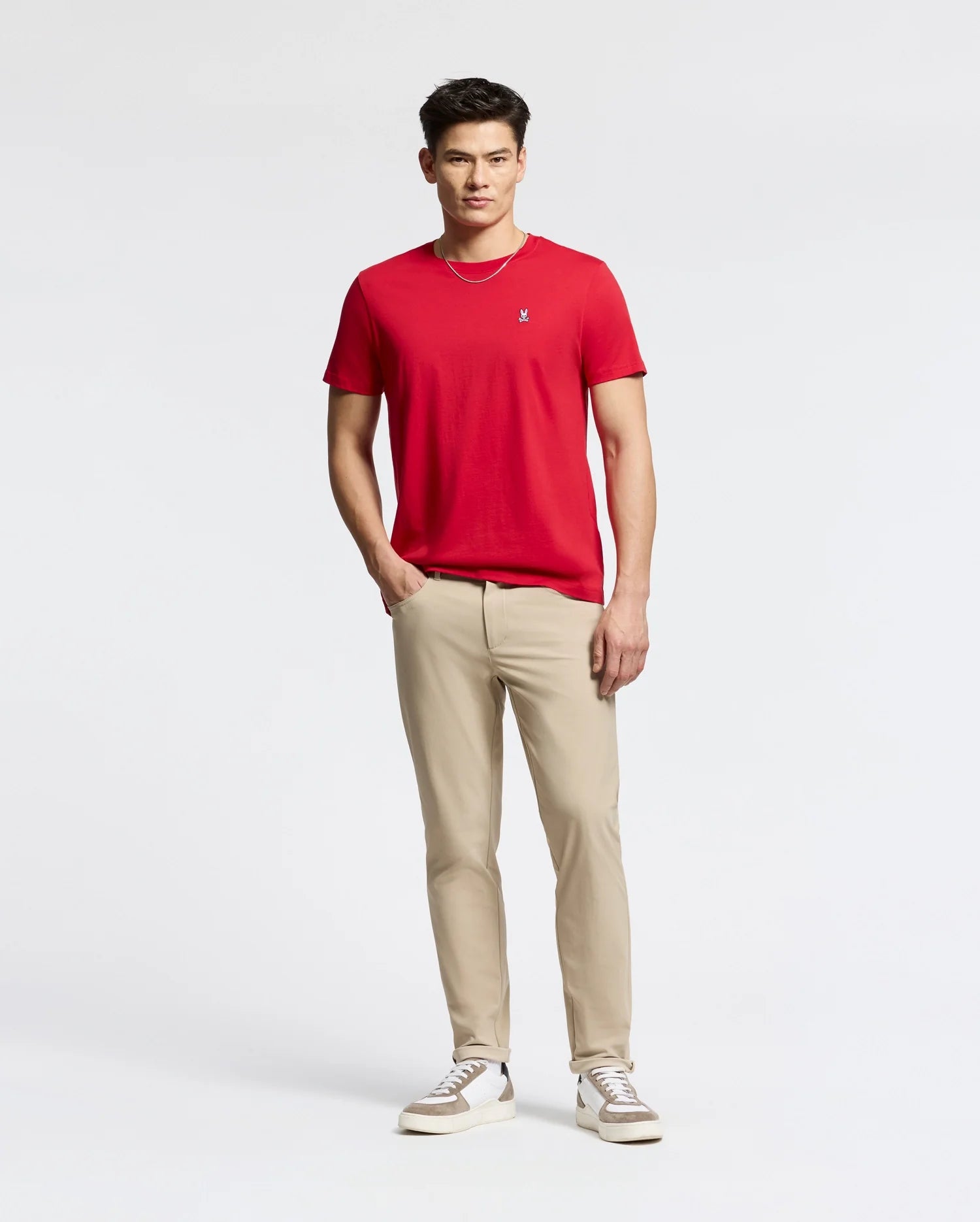 A person with short hair, dressed in a red Psycho Bunny Men's Classic Crew Neck Tee (B6U014CRPC), khaki pants, and white sneakers, stands against a white background. They look at the camera with a neutral expression, showcasing the 100% Pima cotton T-shirt.