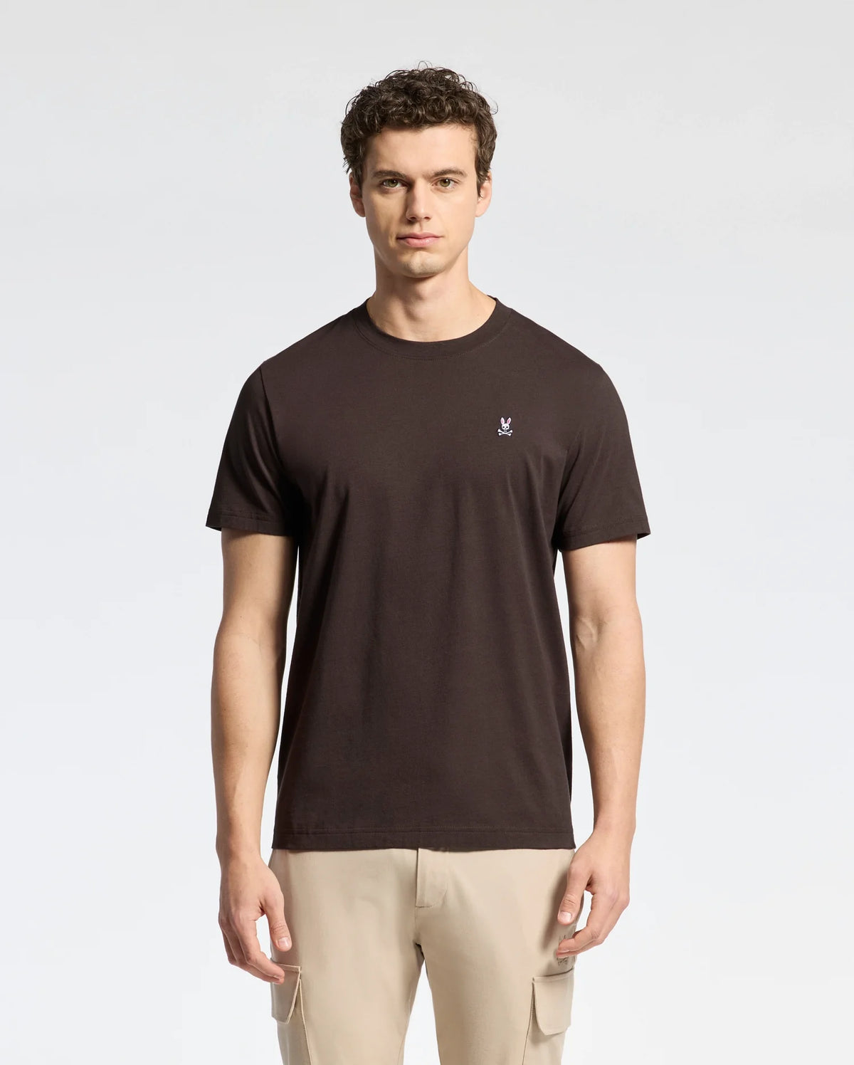 A person with short, curly hair wears a Psycho Bunny MENS CLASSIC CREW NECK TEE - B6U014B200 made of soft Pima cotton jersey and light-colored pants, standing against a neutral background.
