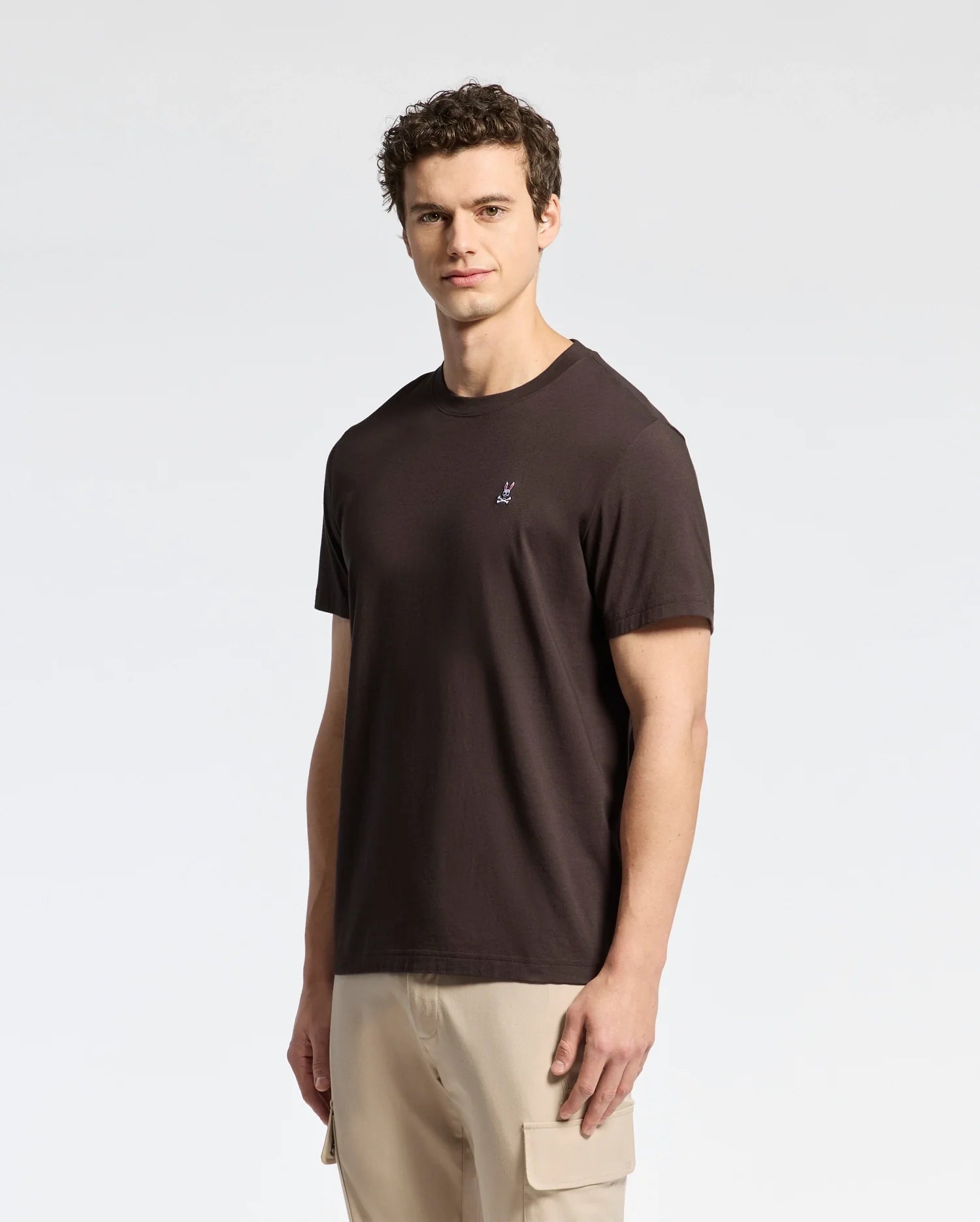 A person with short curly hair wears a Psycho Bunny dark brown MENS CLASSIC CREW NECK TEE - B6U014B200, made from Pima cotton jersey, featuring a chest emblem, and beige cargo pants against a white background.
