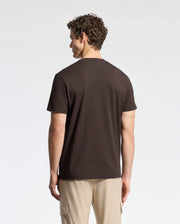 A person with curly hair is wearing a Psycho Bunny MENS CLASSIC CREW NECK TEE - B6U014B200 in dark Pima cotton jersey and beige cargo pants, standing and facing away against a plain white background.