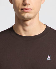 A person wears the Psycho Bunny MENS CLASSIC CREW NECK TEE - B6U014B200 in dark Pima cotton, displaying a white bunny and crossbones logo. The image captures the lower half of the face and upper torso against a muted, vibrant backdrop.