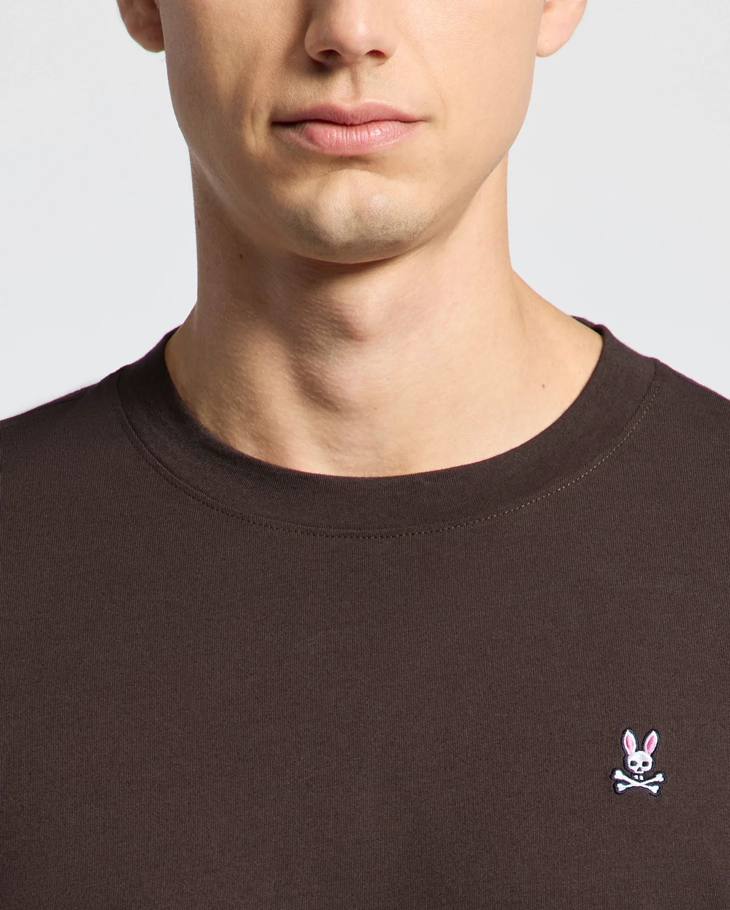 A person wears the Psycho Bunny MENS CLASSIC CREW NECK TEE - B6U014B200 in dark Pima cotton, displaying a white bunny and crossbones logo. The image captures the lower half of the face and upper torso against a muted, vibrant backdrop.