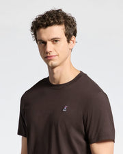 A man with curly hair wears the Psycho Bunny MENS CLASSIC CREW NECK TEE - B6U014B200 in a plain dark shade, gazing at the camera against a light gray backdrop. His soft Pima cotton jersey tee features a small embroidered logo.