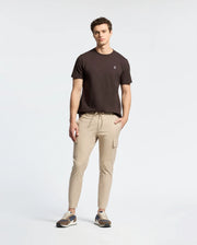A man stands with hands by his sides, wearing a Psycho Bunny Mens Classic Crew Neck Tee in black paired with beige cargo joggers and sneakers against a neutral background.