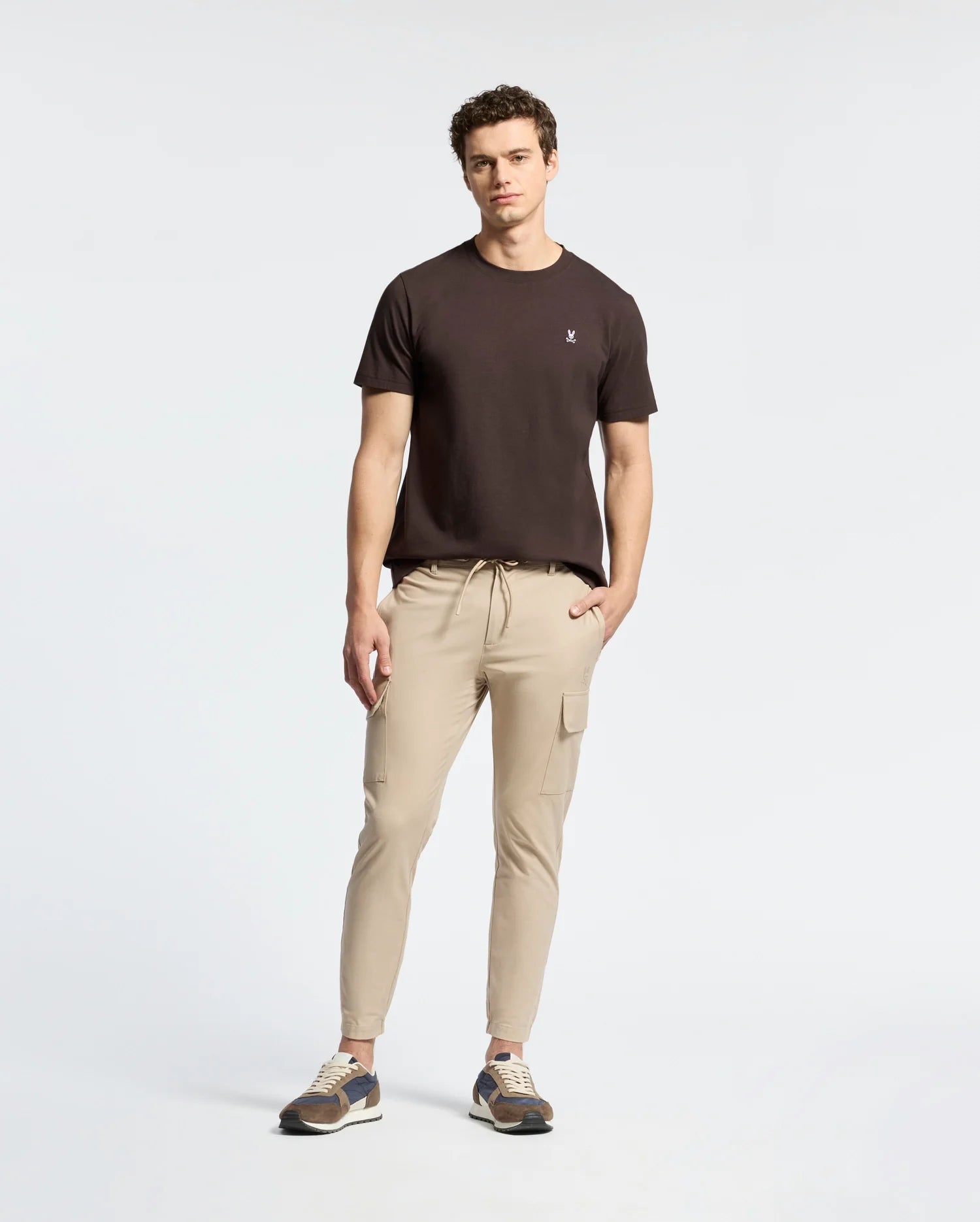 A man stands with hands by his sides, wearing a Psycho Bunny Mens Classic Crew Neck Tee in black paired with beige cargo joggers and sneakers against a neutral background.