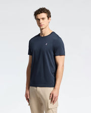 A man with curly hair stands against a white background wearing Psycho Bunny's MENS CLASSIC CREW NECK TEE - B6U014CRPC, made from 100% Pima cotton with an embroidered chest logo. The dark blue T-shirt pairs perfectly with his beige pants featuring a side pocket.