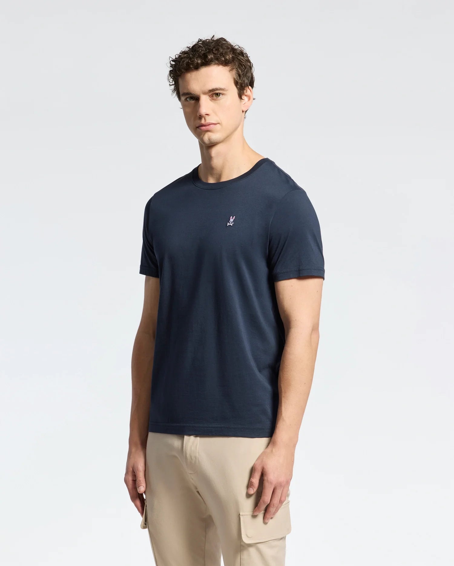 A man with curly hair stands against a white background wearing Psycho Bunny's MENS CLASSIC CREW NECK TEE - B6U014CRPC, made from 100% Pima cotton with an embroidered chest logo. The dark blue T-shirt pairs perfectly with his beige pants featuring a side pocket.