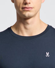 A person is wearing Psycho Bunny's MENS CLASSIC CREW NECK TEE - B6U014CRPC, made from 100% Pima cotton. This tee in a dark blue shade features a small embroidered pink rabbit and crossbones logo on the chest, against a plain light background with their face partially visible.