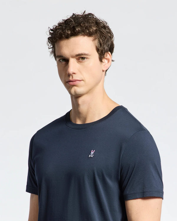 A man with short curly hair stands against a plain light background, wearing a Psycho Bunny navy blue MENS CLASSIC CREW NECK TEE - B6U014CRPC featuring taped seams and an embroidered rabbit logo. Crafted from 100% Pima cotton, it offers style and comfort.