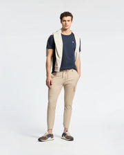 A man stands against a plain background, wearing a Psycho Bunny MENS CLASSIC CREW NECK TEE (B6U014CRPC) in navy blue, made from 100% Pima cotton. He pairs it with beige cargo pants and sneakers. A light grey sweater is draped over his shoulders, with his hands in pockets and short curly hair.
