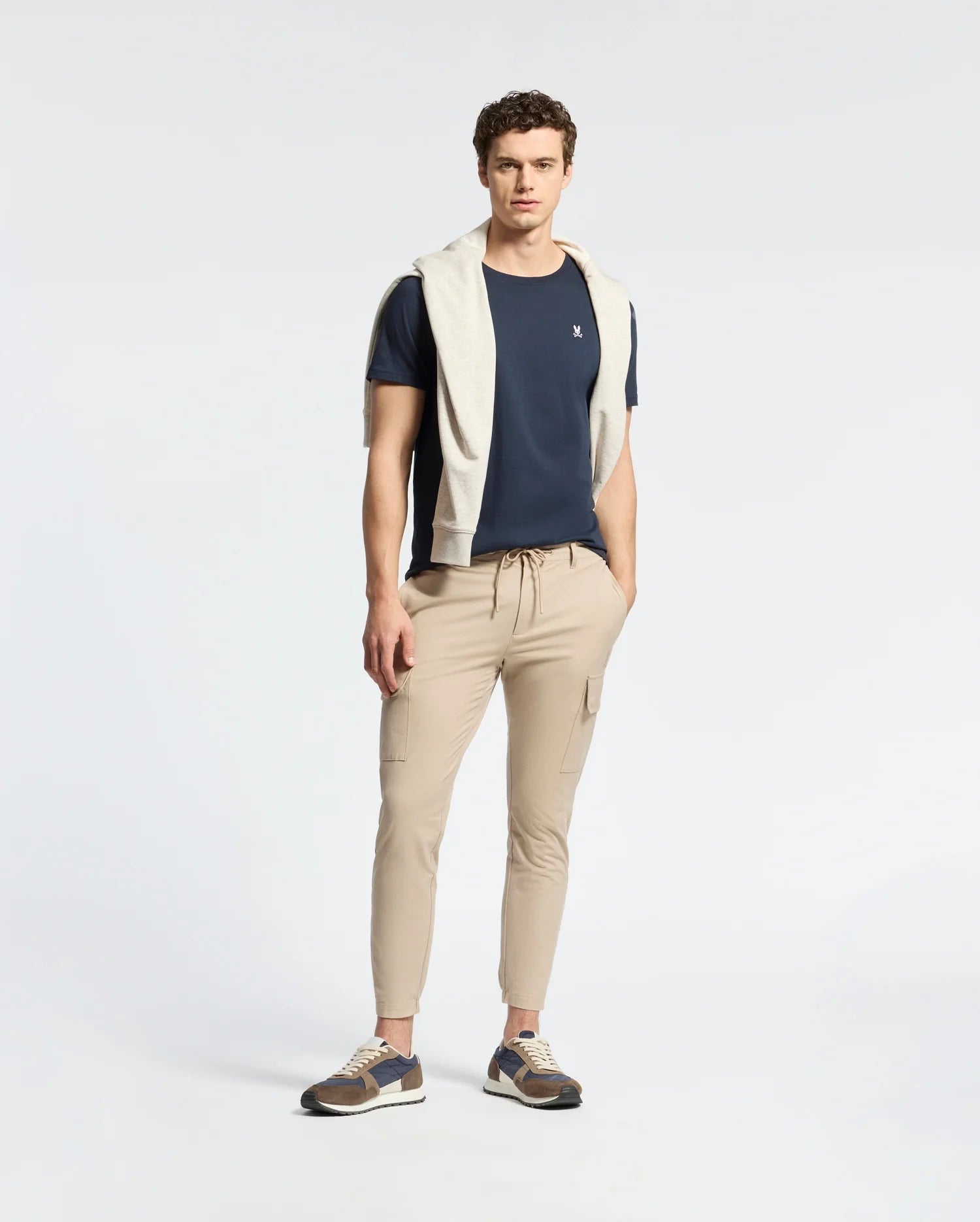 A man stands against a plain background, wearing a Psycho Bunny MENS CLASSIC CREW NECK TEE (B6U014CRPC) in navy blue, made from 100% Pima cotton. He pairs it with beige cargo pants and sneakers. A light grey sweater is draped over his shoulders, with his hands in pockets and short curly hair.