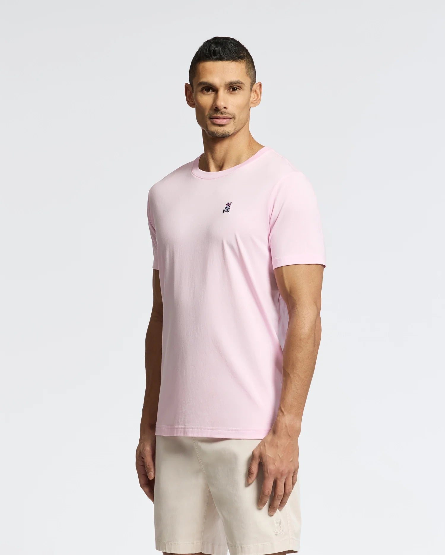 A man stands against a plain backdrop, wearing a Psycho Bunny light pink MENS CLASSIC CREW NECK TEE made of pima cotton with a small chest logo and beige shorts. He has short hair and a neutral expression, showcasing the versatile style.