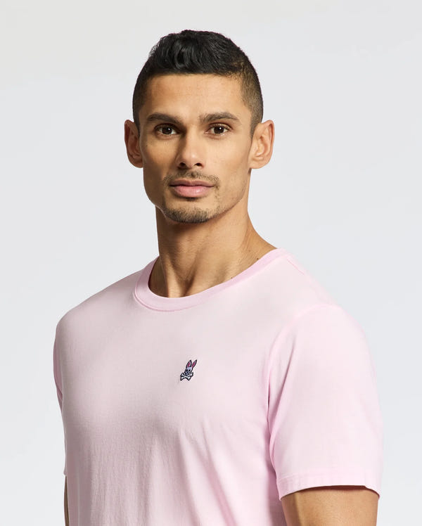 A man with short black hair poses against a plain background, wearing the versatile Psycho Bunny MENS CLASSIC CREW NECK TEE in light pink, made from soft pima cotton and featuring a small embroidered emblem.