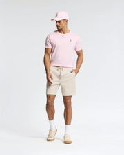 A man models the versatile pink Psycho Bunny MENS CLASSIC CREW NECK TEE - B6U014CRPC, paired with a matching pink cap, beige shorts, white socks, and beige sneakers. Made from soft pima cotton, he stands against a plain white background with his hands in his pockets, gazing to the side.
