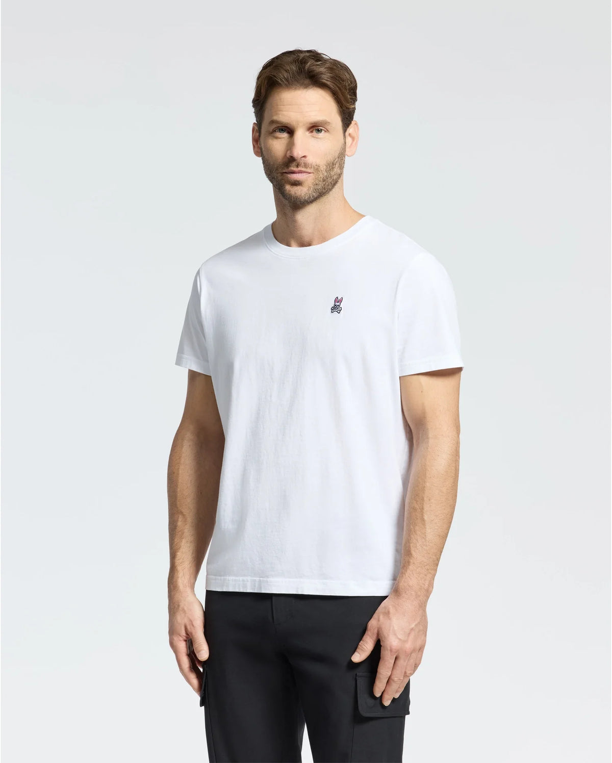 A man with short hair and a beard wears the Psycho Bunny MENS CLASSIC CREW NECK TEE - B6U014CRPC in plain white Pima cotton, with a small embroidered rabbit logo on the left chest. Paired with black pants, he stands against a light gray backdrop, highlighting this men's wardrobe staple.
