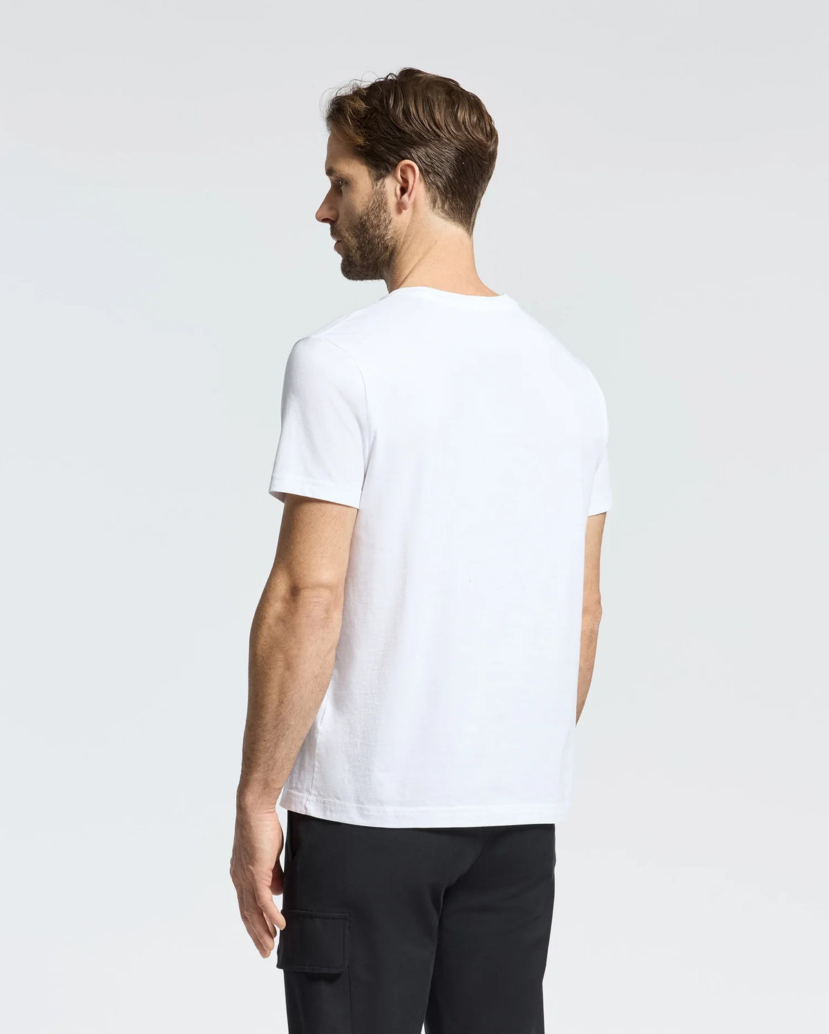 A short-haired man faces away from the camera wearing a Psycho Bunny MENS CLASSIC CREW NECK TEE - B6U014CRPC. The plain white 100% Pima cotton tee with taped seams pairs effortlessly with black pants against a soft gray and white gradient backdrop.