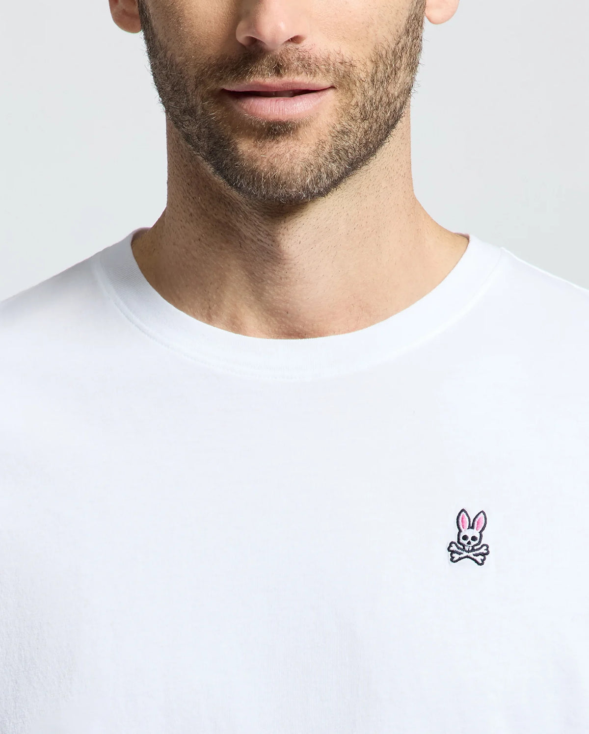 A man wearing a Psycho Bunny men's classic crew neck tee, B6U014CRPC, made of 100% Pima cotton with a small pink bunny and crossbones logo on the chest.