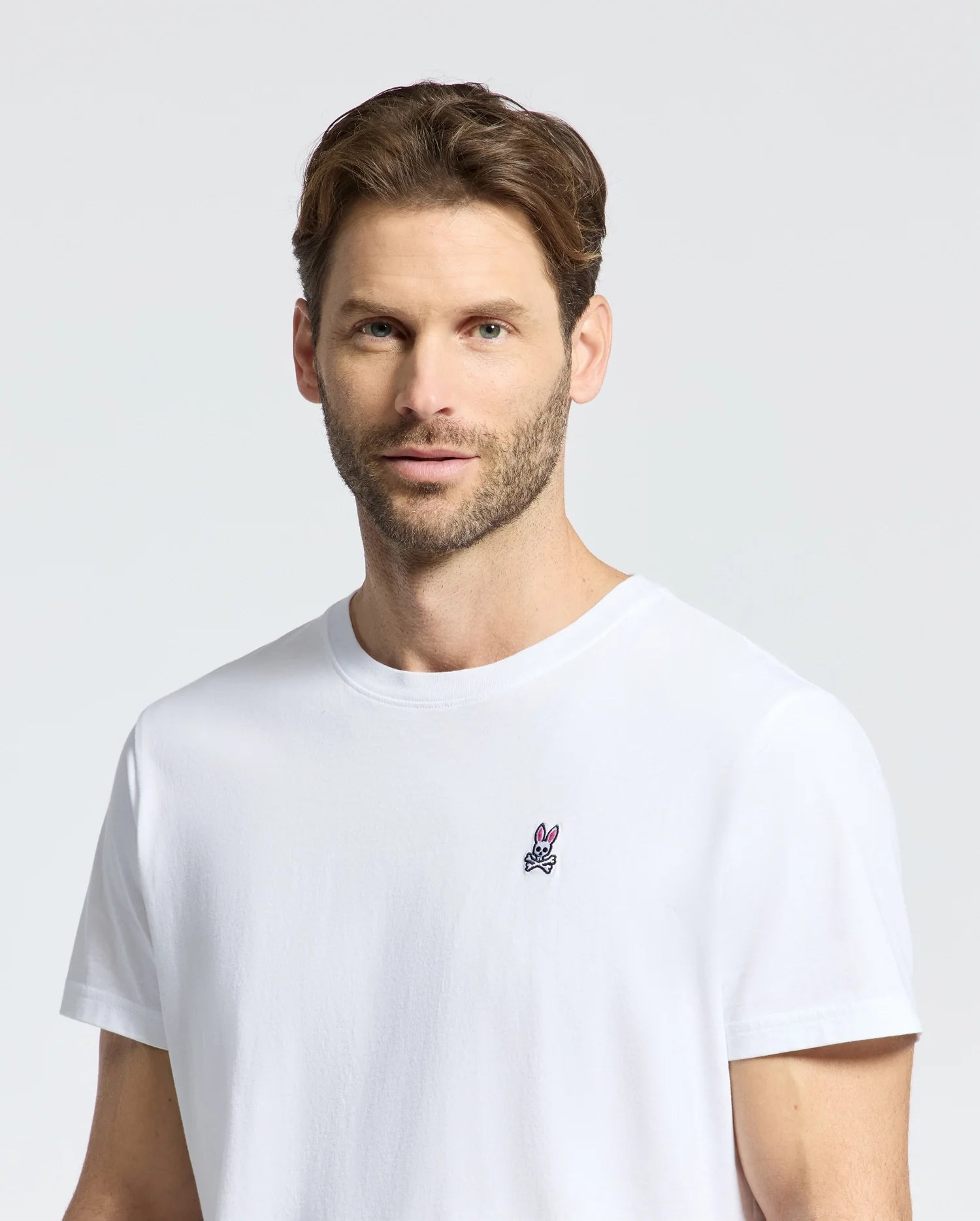 A man with short brown hair and a beard sports the MENS CLASSIC CREW NECK TEE by Psycho Bunny. This plain white Pima cotton t-shirt features a small embroidered bunny logo on the left chest. The background is light gray.