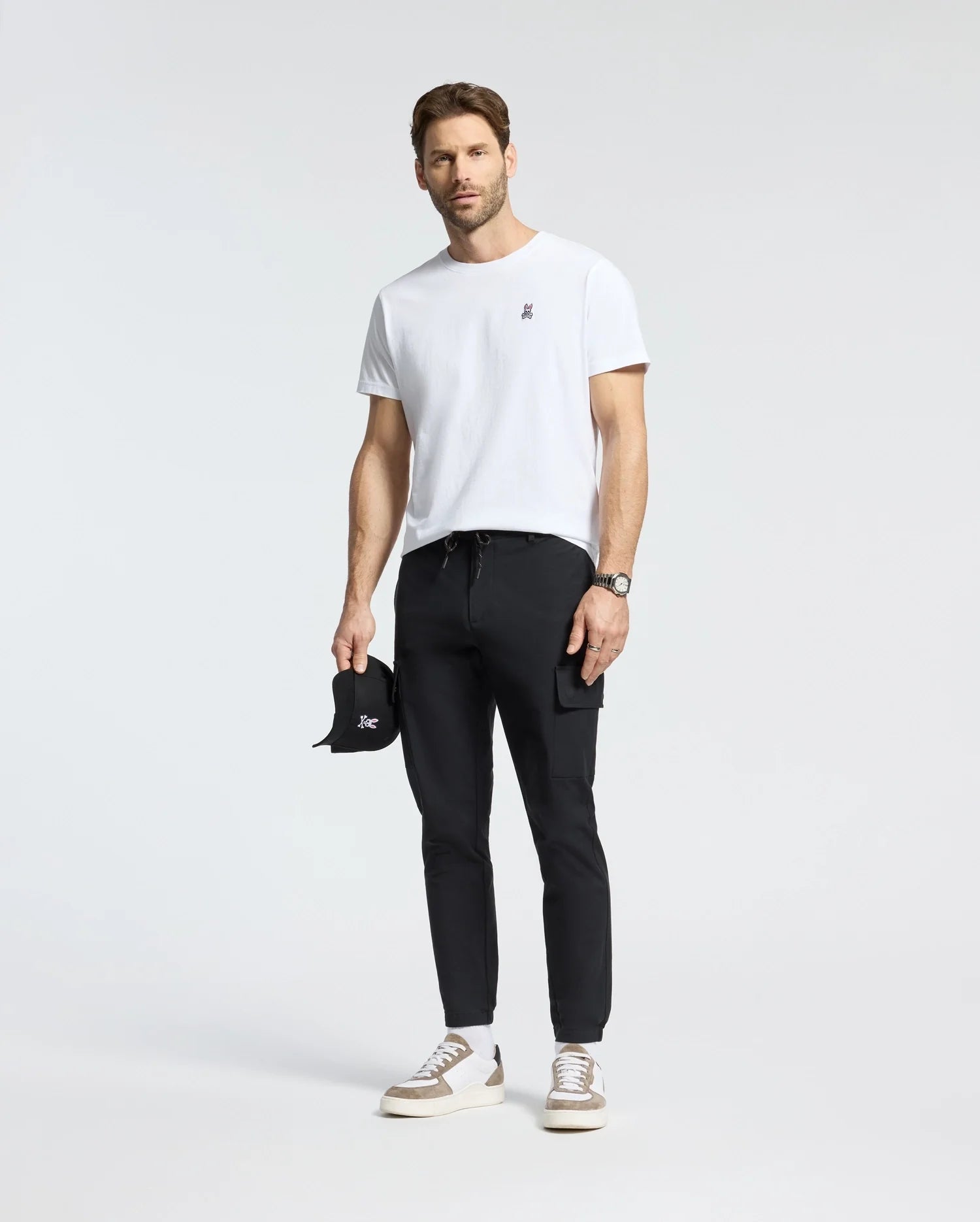 A man is wearing the Psycho Bunny MENS CLASSIC CREW NECK TEE - B6U014CRPC made from 100% Pima cotton, paired with black pants, gray and white sneakers. He holds a black cap and wears a wristwatch, all set against a plain white background that highlights the tee's sleek taped seams.