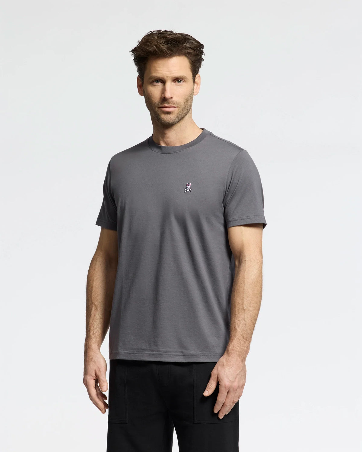 A man with short brown hair and facial hair is wearing a versatile Psycho Bunny MENS CLASSIC CREW NECK TEE (B6U014F200) in gray, crafted from soft pima cotton with a small logo on the chest. He pairs it with black pants and stands against a plain white background, gazing neutrally at the camera.
