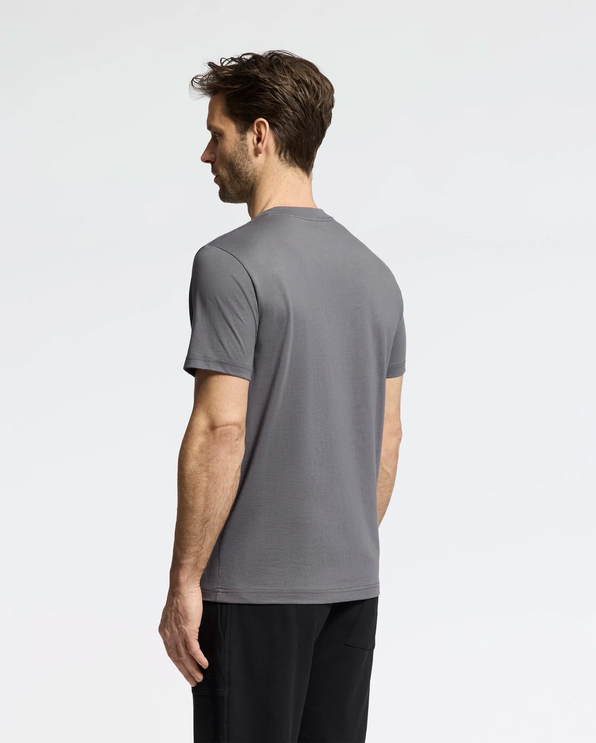 A man with short hair, dressed in a Psycho Bunny MENS CLASSIC CREW NECK TEE in gray and black pants, stands sideways on a plain white background, looking to the left.