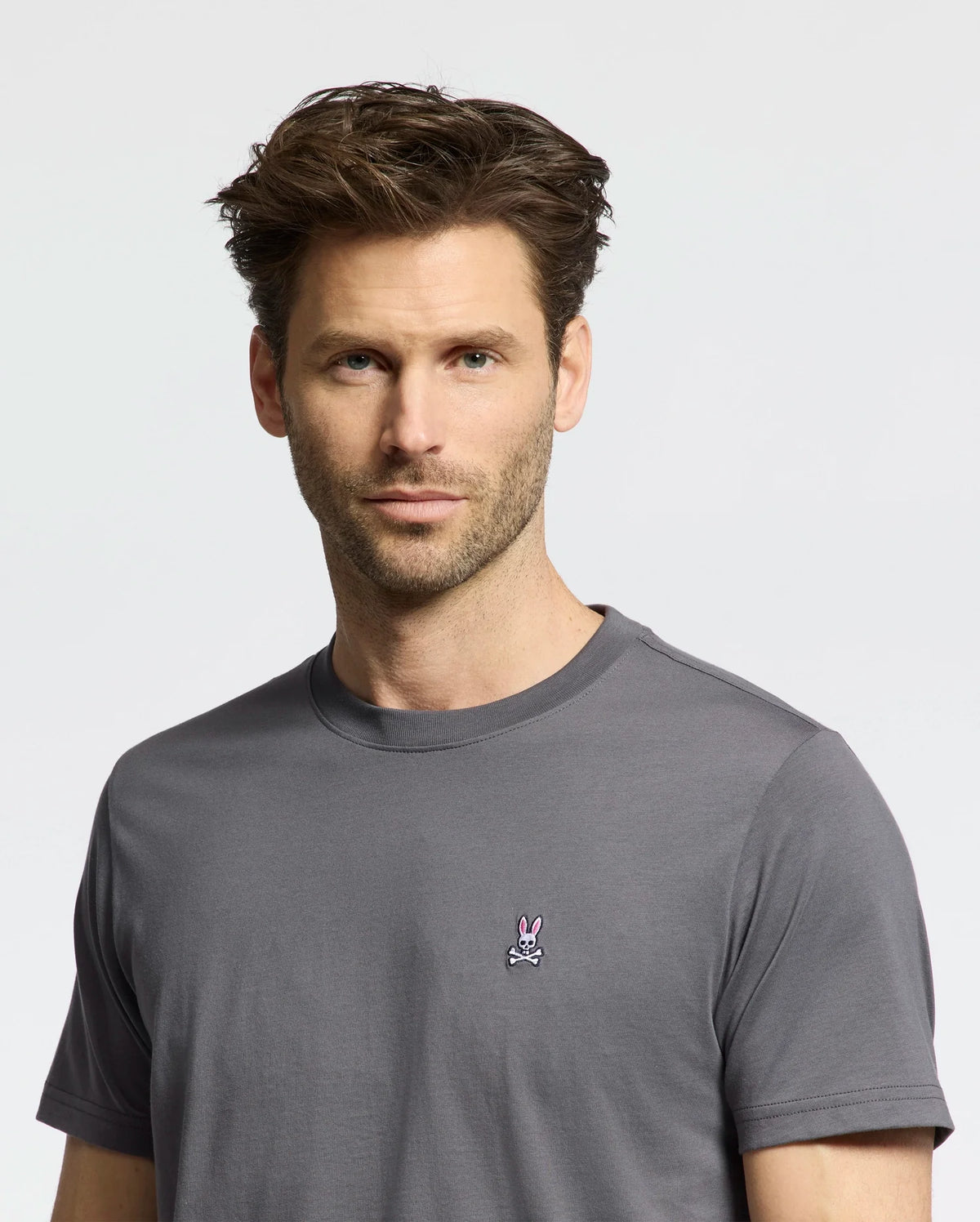 A person with short brown hair wears a Psycho Bunny MENS CLASSIC CREW NECK TEE in gray, showcasing a small embroidered rabbit logo. The background is plain and light-colored.