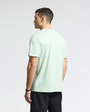 A person with short hair wears the Psycho Bunny MENS CLASSIC CREW NECK TEE - B6U014F200, made of pale green Pima cotton with an embroidered Bunny logo. They're turned slightly away from the camera, and it pairs seamlessly with black pants against a plain white backdrop.
