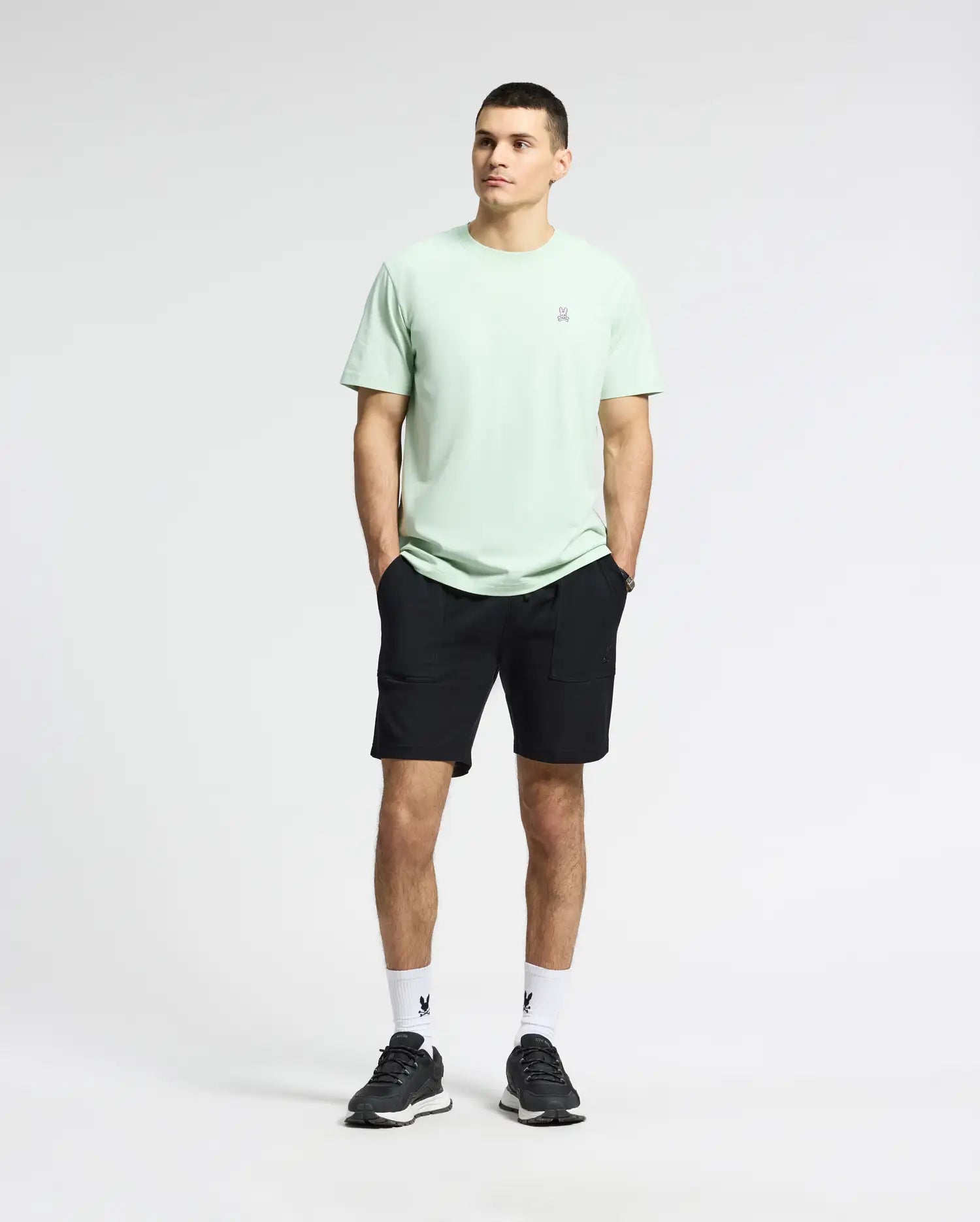 A person stands against a plain background in a light green MENS CLASSIC CREW NECK TEE - B6U014F200 by Psycho Bunny, paired with black shorts, white socks, and black sneakers. They have short hair and their hands in pockets, the embroidered Bunny logo visible as they look slightly left.